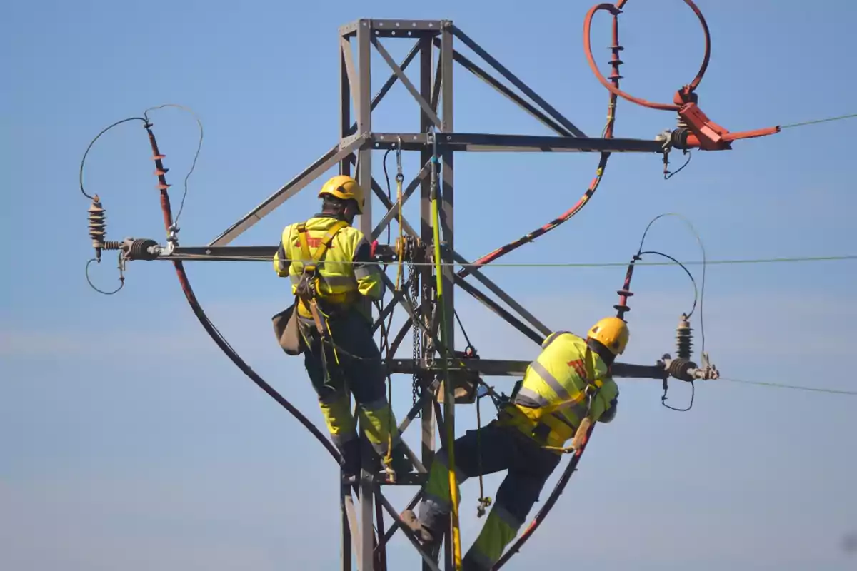 Dos trabajadores con equipo de seguridad realizan labores de mantenimiento en una torre de alta tensión bajo un cielo despejado
