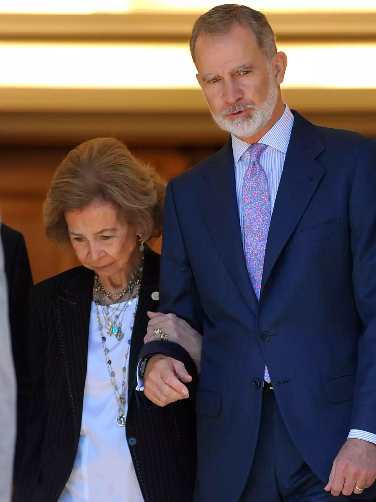 Felipe VI con traje azul ayuda a la reina Sofía que lleva un conjunto oscuro y varios collares Felipe VI con traje azul ayuda a la reina Sofía que lleva un conjunto oscuro y varios collares