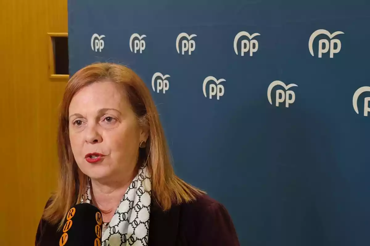 Mujer hablando frente a un micrófono con fondo azul que tiene varios logotipos del Partido Popular