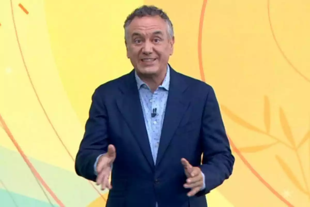 Plano medio de Roberto Brasero vestido con traje azul y camisa clara hablando frente a un fondo amarillo con líneas y formas abstractas. Plano medio de Roberto Brasero vestido con traje azul y camisa clara hablando frente a un fondo amarillo con líneas y formas abstractas.