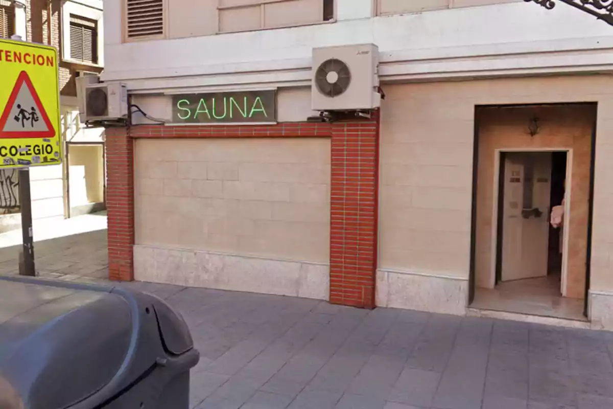 Fachada de un edificio con un letrero de neón que dice sauna, un cartel amarillo de advertencia de colegio y una puerta abierta Fachada de un edificio con un letrero de neón que dice sauna, un cartel amarillo de advertencia de colegio y una puerta abierta