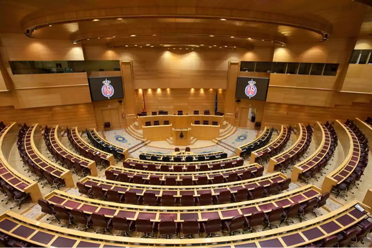 Vista del hemiciclo del Senado de España con asientos vacíos y dos pantallas mostrando el escudo oficial Vista del hemiciclo del Senado de España con asientos vacíos y dos pantallas mostrando el escudo oficial
