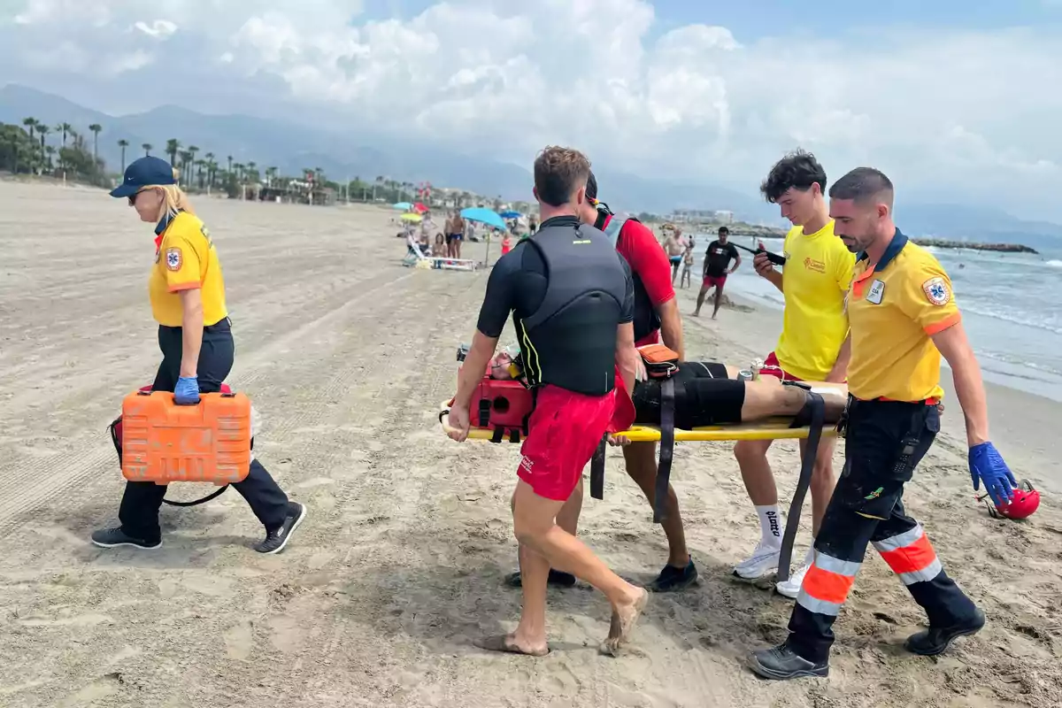 Socorristas y personal de emergencia trasladan a una persona herida en camilla por la playa mientras una rescatista lleva un maletín naranja de primeros auxilios Socorristas y personal de emergencia trasladan a una persona herida en camilla por la playa mientras una rescatista lleva un maletín naranja de primeros auxilios