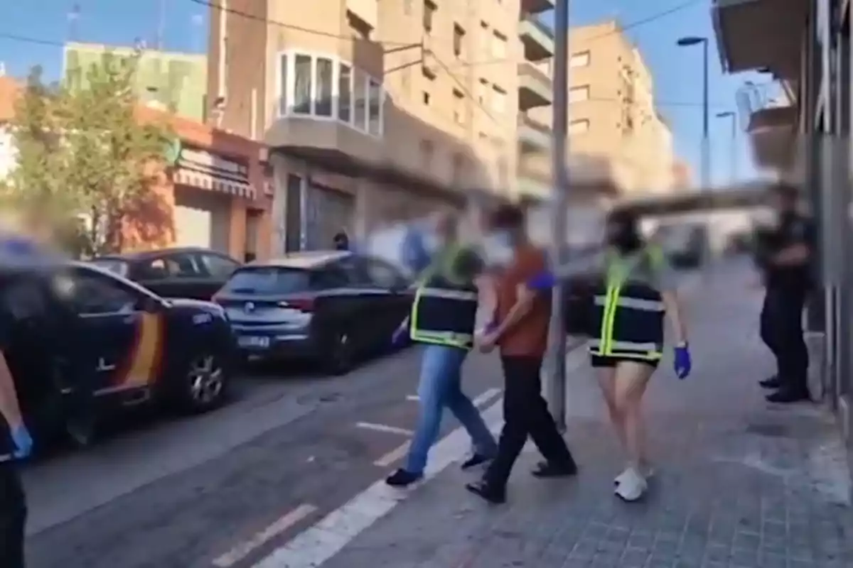 Varias personas, entre ellas dos agentes con chalecos reflectantes, escoltan a un hombre esposado por una acera en una calle urbana con coches y edificios al fondo.