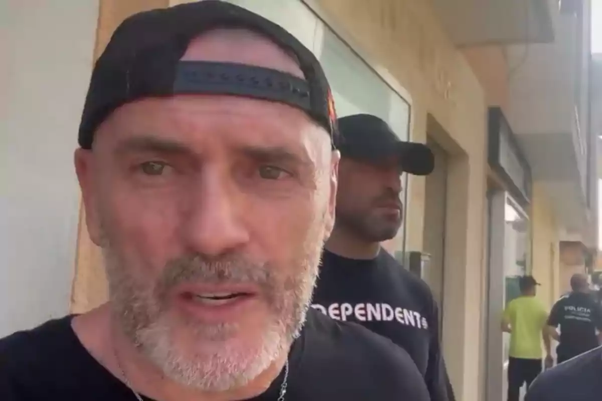 Hombre de barba y gorra negra al revés en primer plano con otra persona y varias personas al fondo en una calle Hombre de barba y gorra negra al revés en primer plano con otra persona y varias personas al fondo en una calle