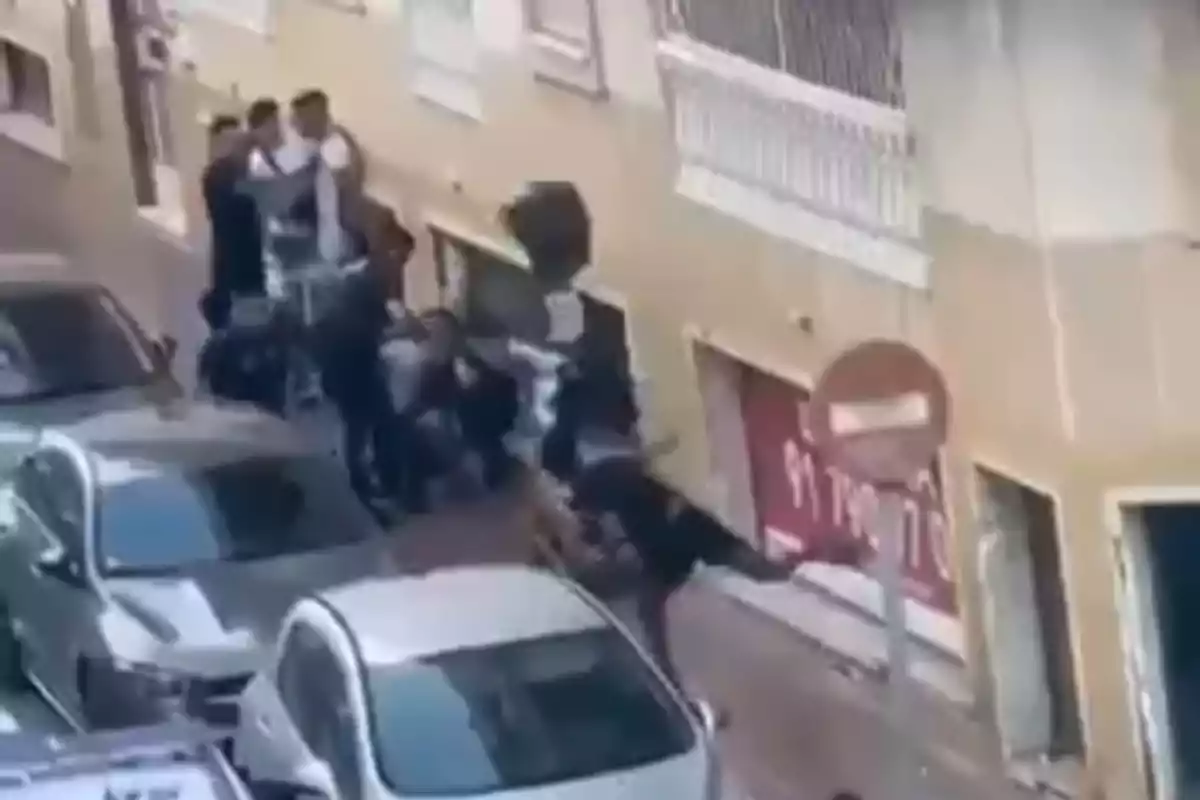 Varias personas se encuentran reunidas en una acera junto a una fila de autos estacionados en una calle estrecha frente a un edificio beige