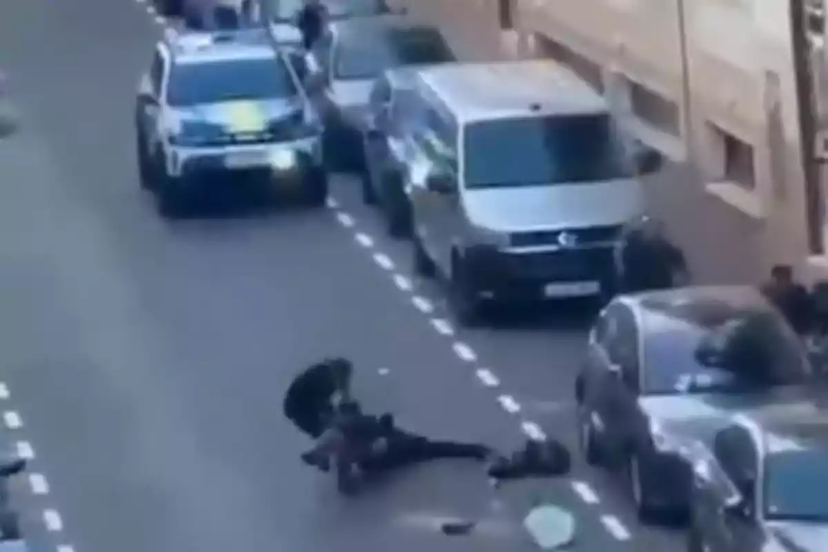 Un policía reduce a una persona en el suelo en medio de una calle con varios autos estacionados y una patrulla al fondo Un policía reduce a una persona en el suelo en medio de una calle con varios autos estacionados y una patrulla al fondo