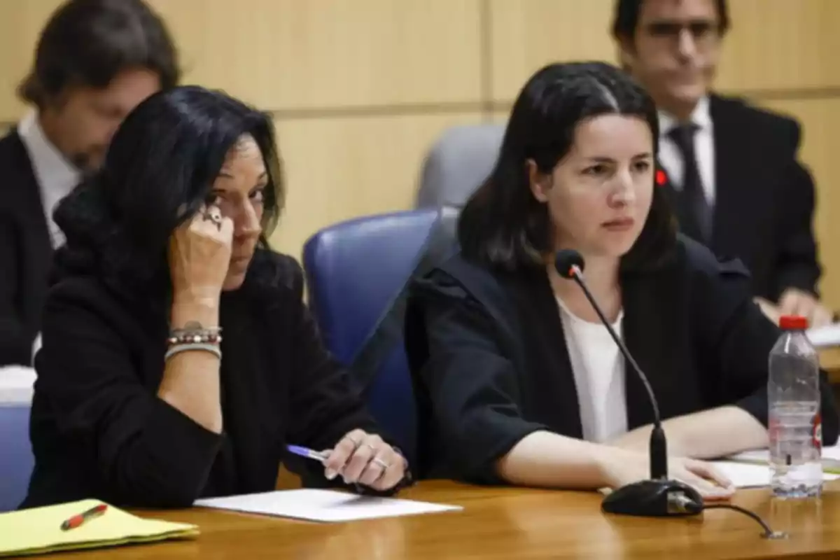 Dos mujeres sentadas en una mesa durante una audiencia o juicio, una de ellas sostiene un bolígrafo y la otra habla frente a un micrófono, ambas con expresión seria y documentos sobre la mesa