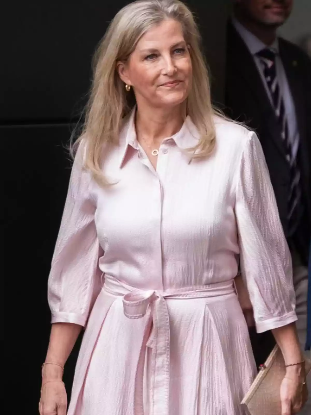 Blonde, long-haired woman wearing a light pink three-quarter sleeve dress with a belt, holding a beige handbag and smiling slightly Blonde, long-haired woman wearing a light pink three-quarter sleeve dress with a belt, holding a beige handbag and smiling slightly