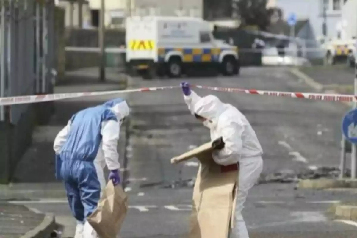 Dos personas con trajes forenses recogen evidencia en una calle acordonada por la policía mientras un vehículo policial se encuentra al fondo Dos personas con trajes forenses recogen evidencia en una calle acordonada por la policía mientras un vehículo policial se encuentra al fondo