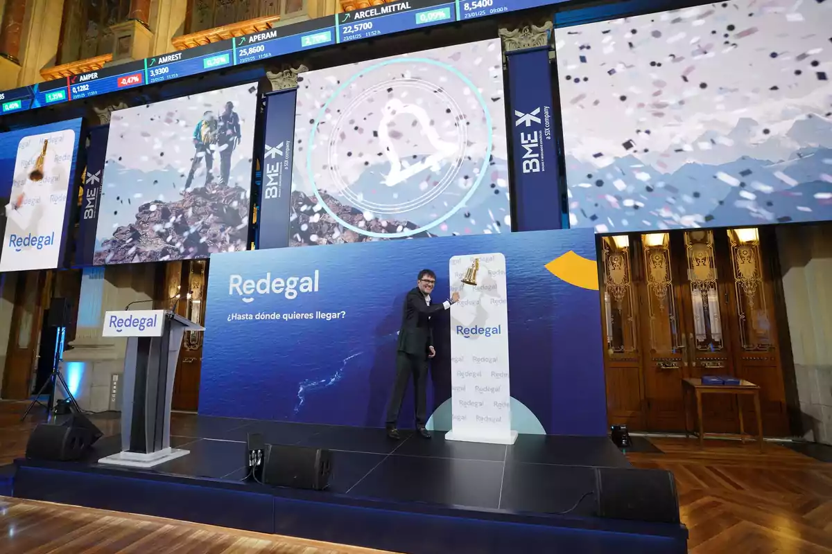 Un hombre toca una campana en un escenario decorado con el logo de Redegal y pantallas grandes, en un evento corporativo en la bolsa de valores.