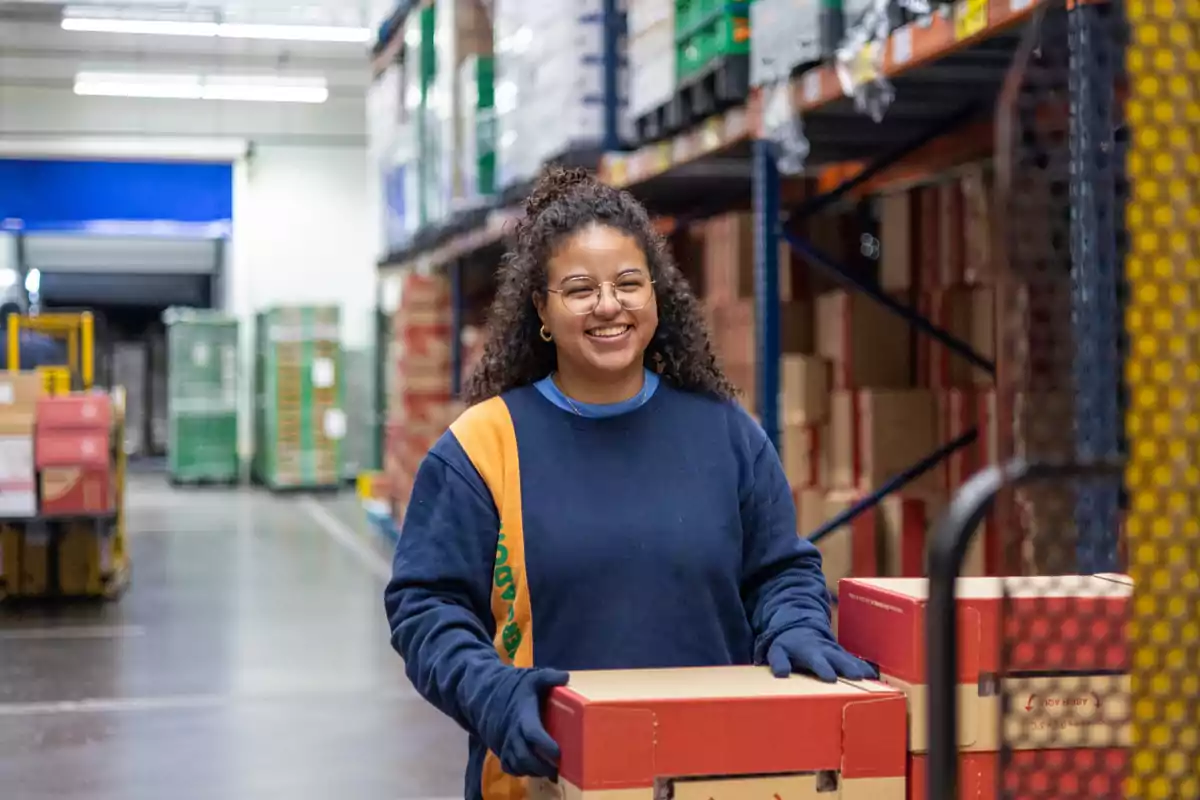 Mujer sonriente con gafas y guantes trabajando en un almacén mientras mueve cajas de cartón Mujer sonriente con gafas y guantes trabajando en un almacén mientras mueve cajas de cartón