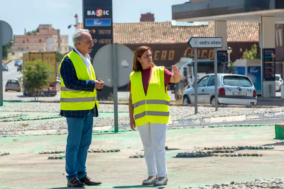 Dos personas con chalecos reflectantes conversan en una zona urbana cerca de una gasolinera y señales de tráfico Dos personas con chalecos reflectantes conversan en una zona urbana cerca de una gasolinera y señales de tráfico