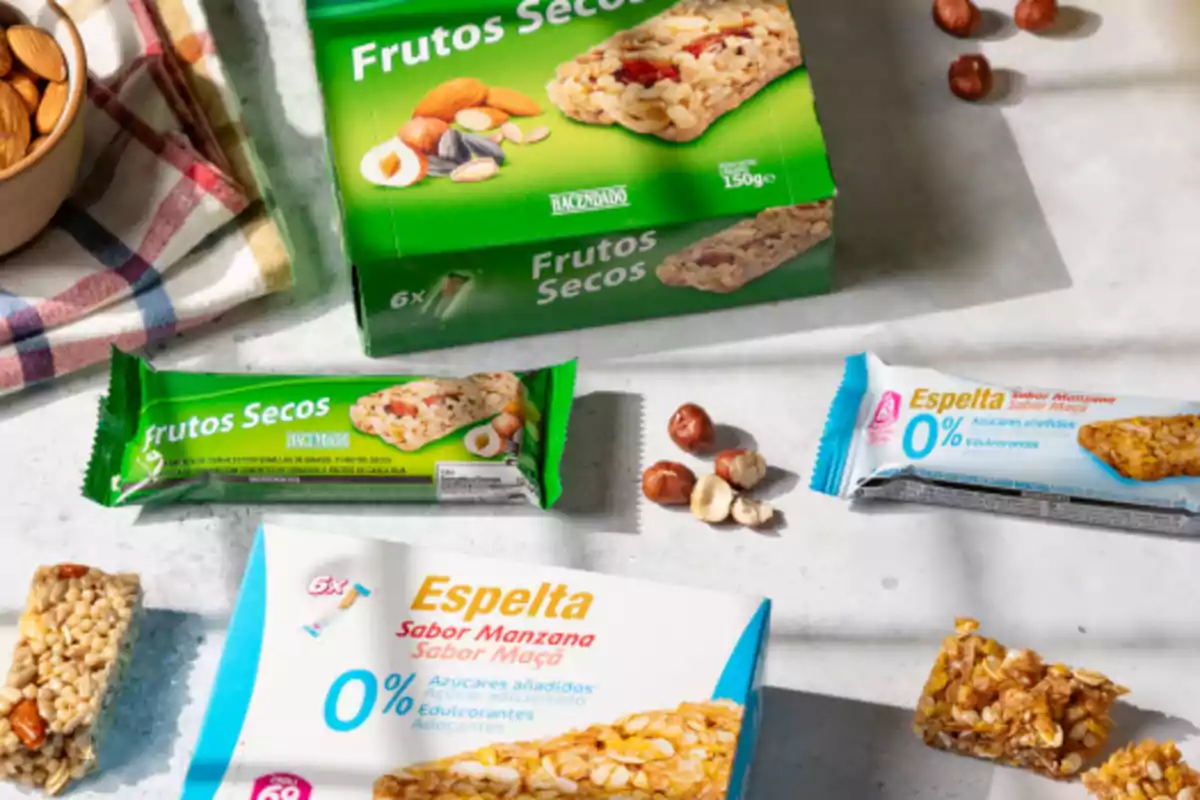 Barras de cereales y frutos secos en diferentes empaques sobre una mesa junto a algunas nueces y almendras Barras de cereales y frutos secos en diferentes empaques sobre una mesa junto a algunas nueces y almendras