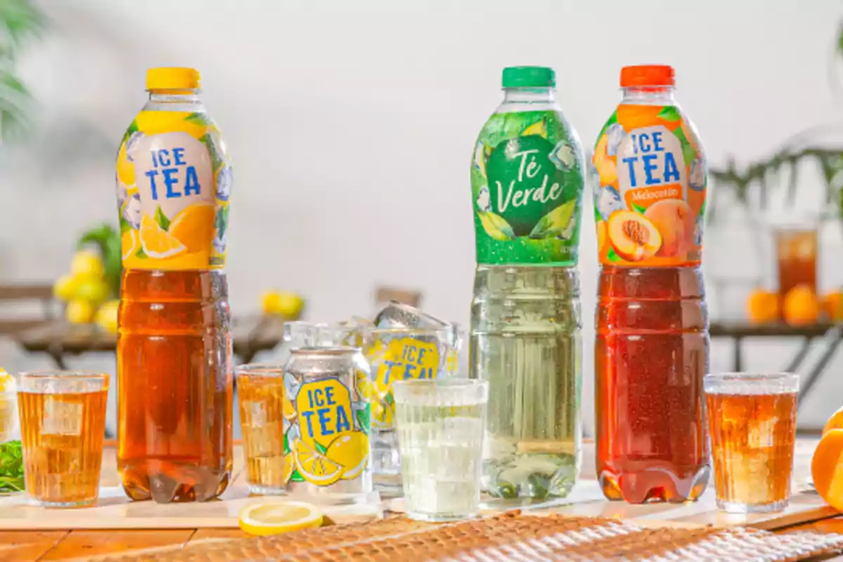 Tres botellas de té helado de diferentes sabores junto a vasos y una lata del mismo producto sobre una mesa, con frutas y bebidas al fondo en un ambiente luminoso Tres botellas de té helado de diferentes sabores junto a vasos y una lata del mismo producto sobre una mesa, con frutas y bebidas al fondo en un ambiente luminoso