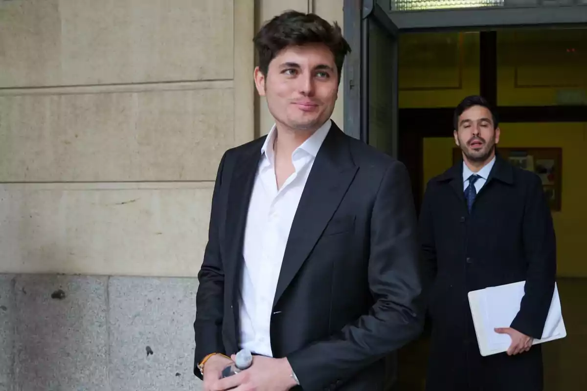 Hombre joven con traje oscuro y camisa blanca de pie frente a un edificio mientras otra persona con abrigo y carpeta sale detrás de él