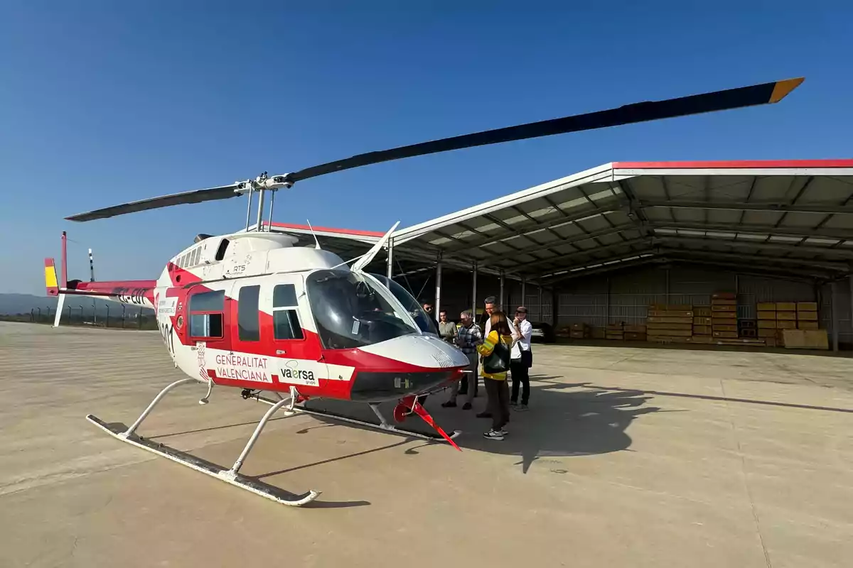 Helicóptero blanco y rojo estacionado en una pista de concreto junto a un hangar con varias personas de pie cerca de la puerta del helicóptero en un día soleado Helicóptero blanco y rojo estacionado en una pista de concreto junto a un hangar con varias personas de pie cerca de la puerta del helicóptero en un día soleado