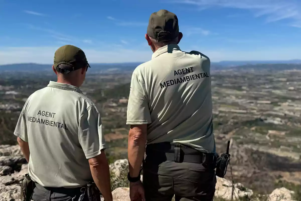Dos agentes medioambientales de espaldas observan un paisaje natural desde lo alto de una montaña