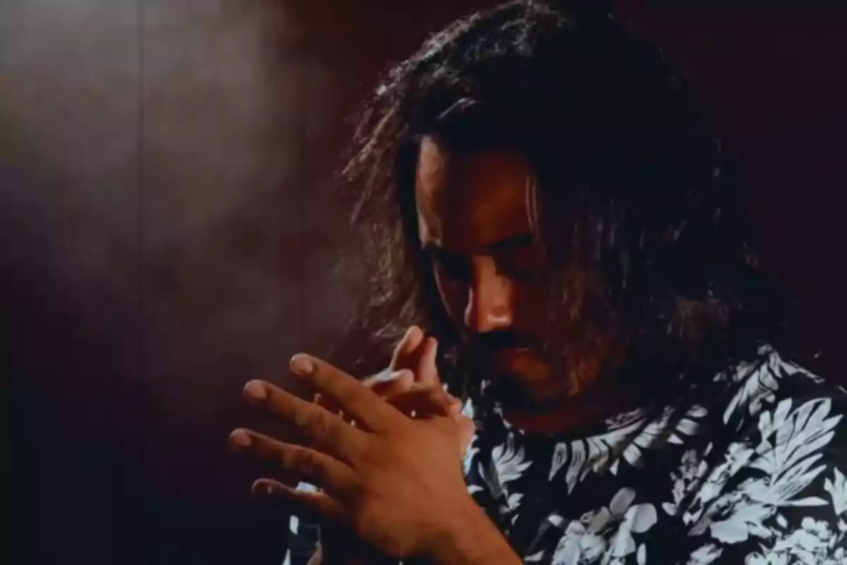 Hombre de cabello largo y barba con expresión pensativa, usando una camiseta de flores y las manos entrelazadas, en un ambiente oscuro
