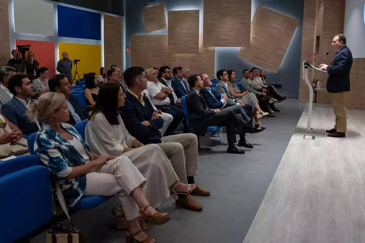 Un hombre de pie da una conferencia frente a un público sentado en una sala moderna con paneles acústicos en las paredes y cámaras grabando el evento. Un hombre de pie da una conferencia frente a un público sentado en una sala moderna con paneles acústicos en las paredes y cámaras grabando el evento.