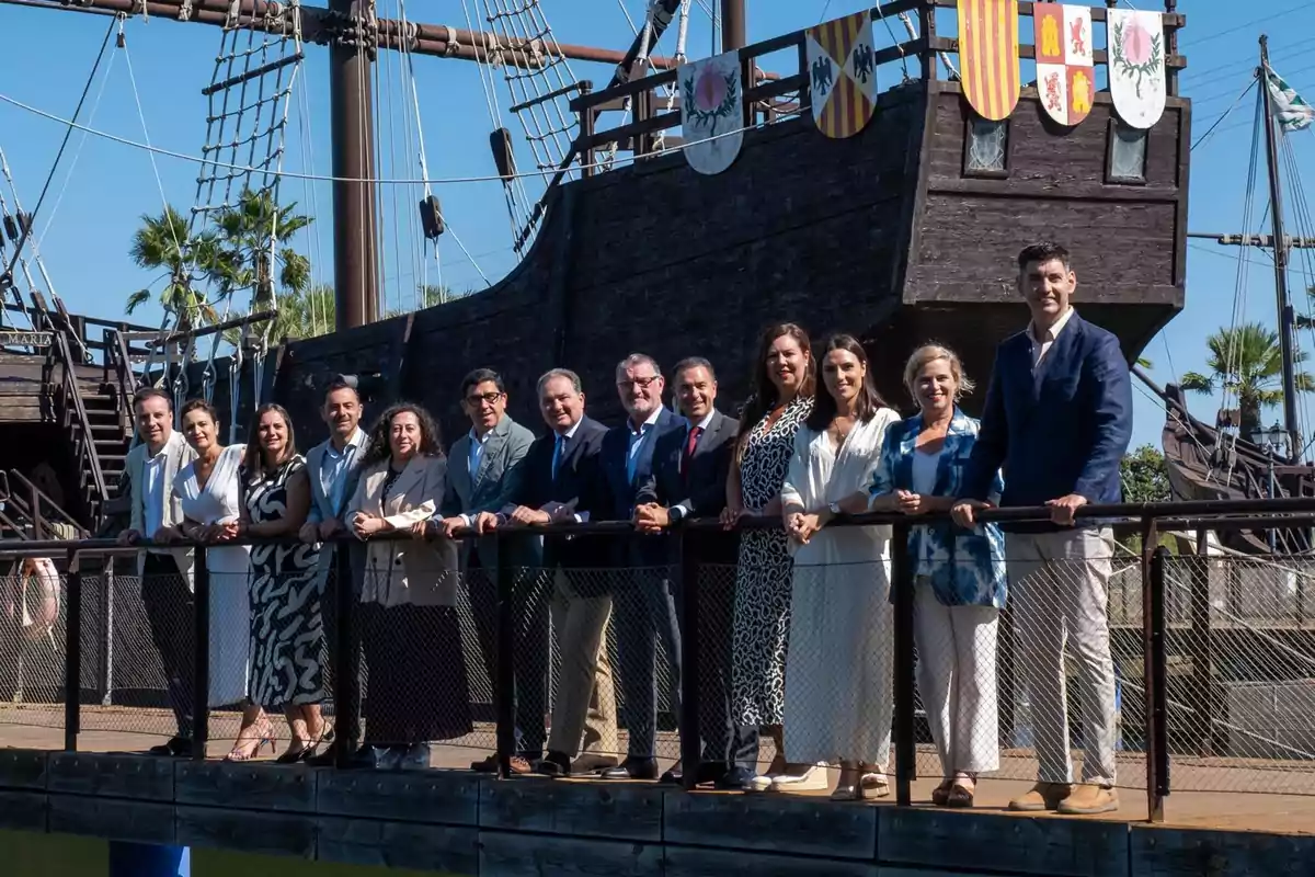 Un grupo de personas posando sobre un puente de madera frente a una réplica de un barco antiguo con escudos en la parte superior y palmeras al fondo en un día soleado. Un grupo de personas posando sobre un puente de madera frente a una réplica de un barco antiguo con escudos en la parte superior y palmeras al fondo en un día soleado.