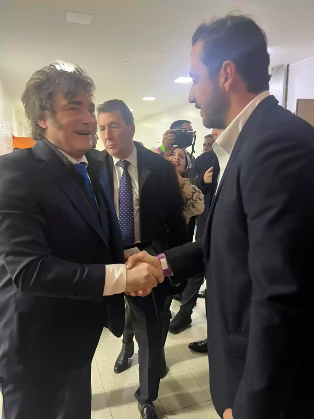 Dos hombres vestidos de traje se dan la mano en un pasillo mientras otras personas los observan y una mujer toma una foto al fondo Dos hombres vestidos de traje se dan la mano en un pasillo mientras otras personas los observan y una mujer toma una foto al fondo