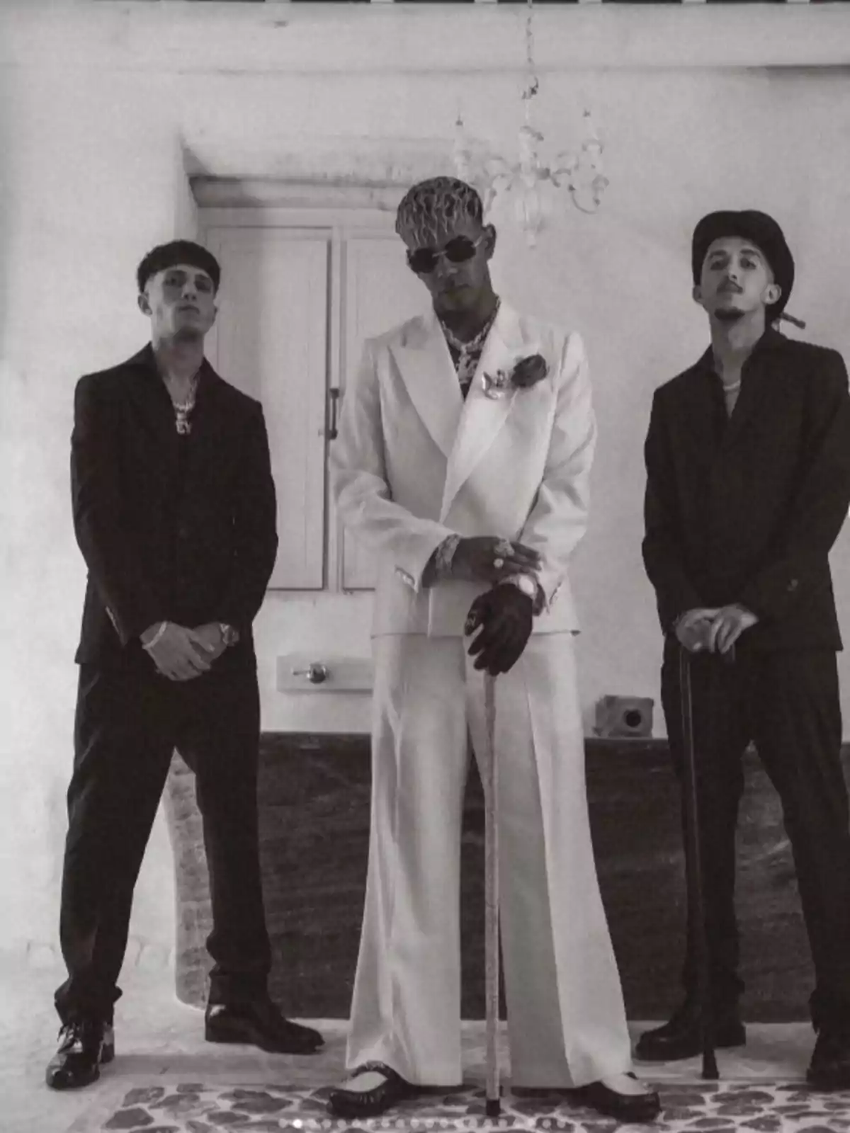 Tres hombres posan en una habitación elegante, dos de ellos vestidos de negro y uno en el centro con un traje blanco, gafas de sol y bastón, todos con expresión seria Tres hombres posan en una habitación elegante, dos de ellos vestidos de negro y uno en el centro con un traje blanco, gafas de sol y bastón, todos con expresión seria