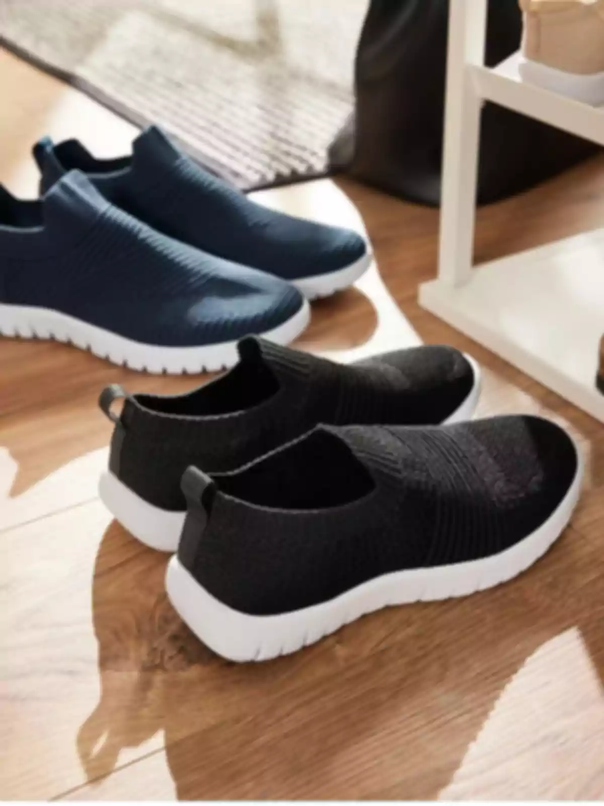 Dos pares de tenis deportivos sin agujetas en colores negro y azul marino con suela blanca sobre un piso de madera Dos pares de tenis deportivos sin agujetas en colores negro y azul marino con suela blanca sobre un piso de madera