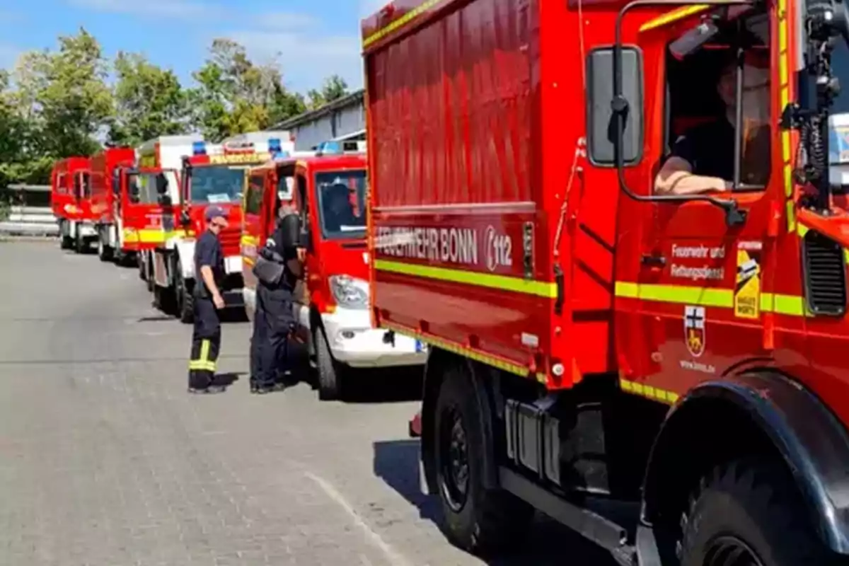 Varios camiones de bomberos rojos alineados en una calle con bomberos de pie junto a los vehículos en un día soleado. Varios camiones de bomberos rojos alineados en una calle con bomberos de pie junto a los vehículos en un día soleado.