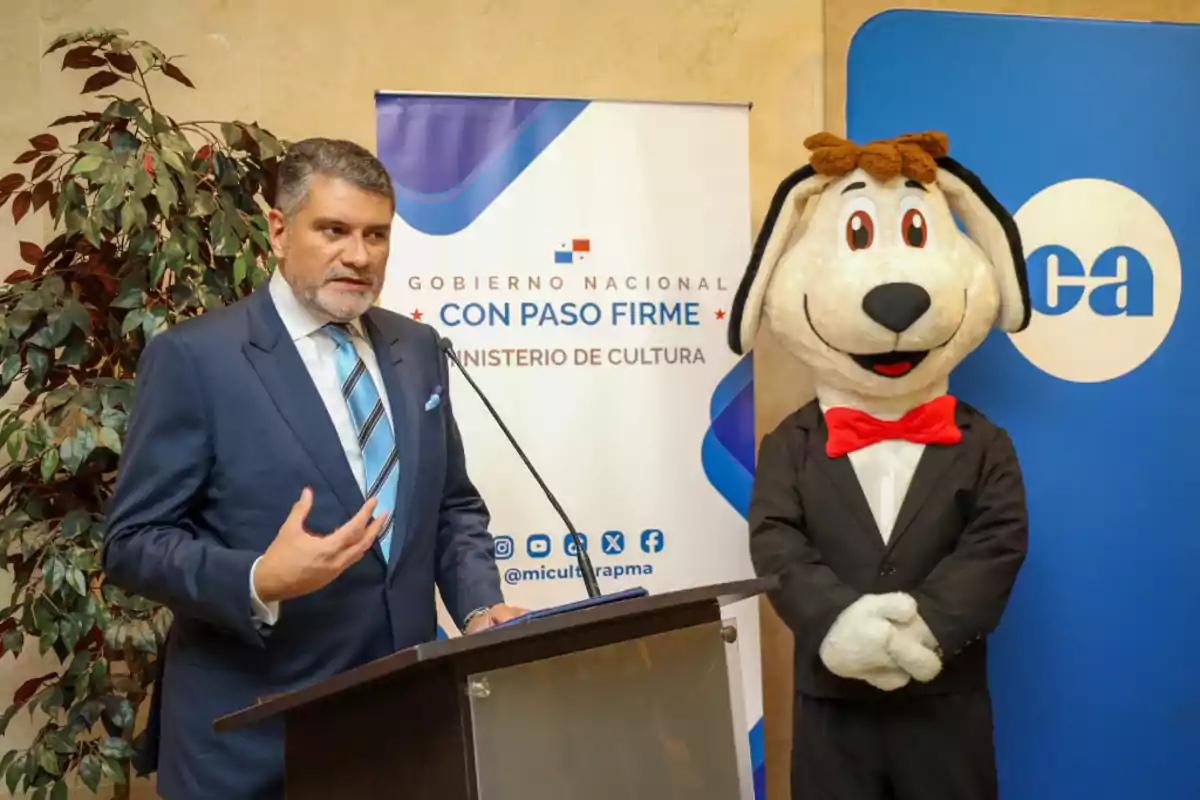 Un hombre de traje habla en un podio mientras una persona disfrazada de perro con esmoquin y moño rojo está de pie a su lado