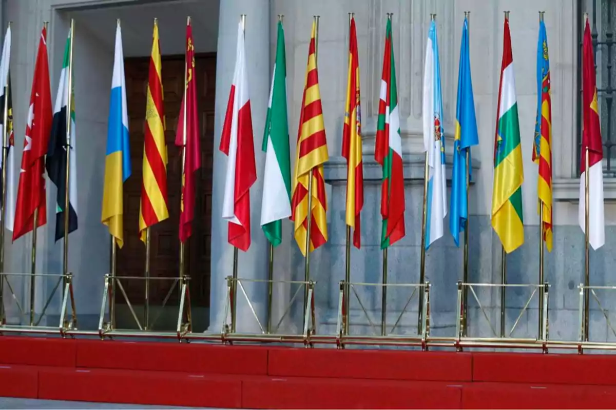 Varias banderas de diferentes comunidades autónomas de España están alineadas sobre una tarima roja frente a un edificio oficial