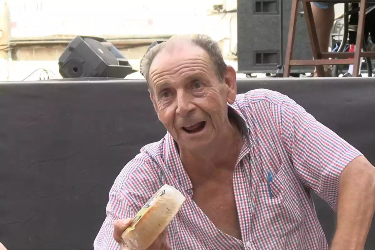 Hombre mayor sonriente con camisa de cuadros sostiene un queso pequeño en la mano durante un evento al aire libre Hombre mayor sonriente con camisa de cuadros sostiene un queso pequeño en la mano durante un evento al aire libre
