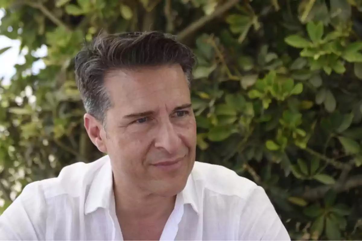 Hombre de cabello oscuro y camisa blanca sentado al aire libre con plantas verdes de fondo Hombre de cabello oscuro y camisa blanca sentado al aire libre con plantas verdes de fondo