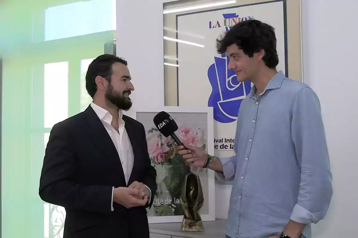Un hombre con barba y traje oscuro es entrevistado por otro hombre con camisa azul que sostiene un micrófono en una sala con cuadros y una escultura sobre una mesa Un hombre con barba y traje oscuro es entrevistado por otro hombre con camisa azul que sostiene un micrófono en una sala con cuadros y una escultura sobre una mesa