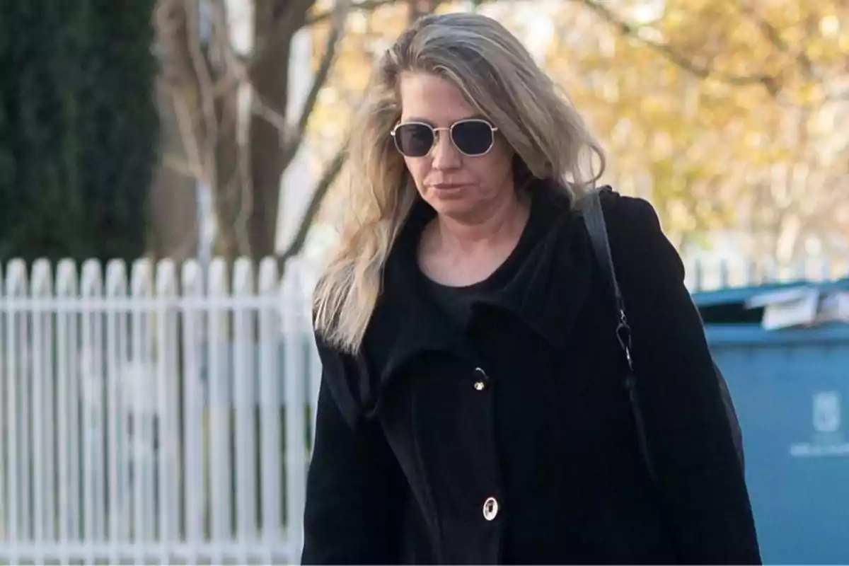 Mujer rubia con gafas de sol y abrigo negro caminando al aire libre frente a una cerca blanca Mujer rubia con gafas de sol y abrigo negro caminando al aire libre frente a una cerca blanca