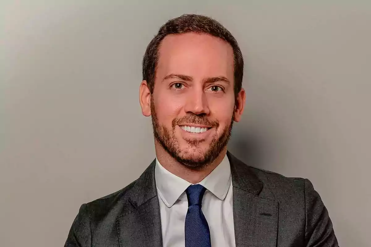 Hombre joven con barba y cabello corto, vestido con traje gris, camisa blanca y corbata azul, sonriendo frente a un fondo gris. Hombre joven con barba y cabello corto, vestido con traje gris, camisa blanca y corbata azul, sonriendo frente a un fondo gris.