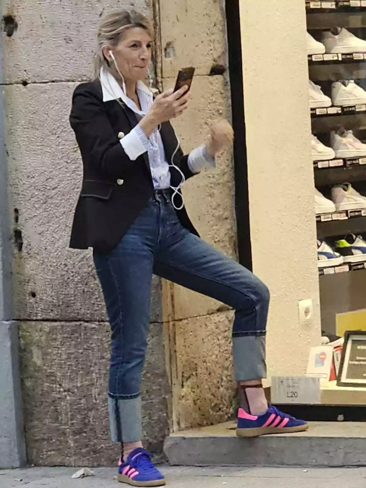 Mujer de pie en la calle usando auriculares y mirando su teléfono móvil frente a una tienda de zapatillas deportivas Mujer de pie en la calle usando auriculares y mirando su teléfono móvil frente a una tienda de zapatillas deportivas