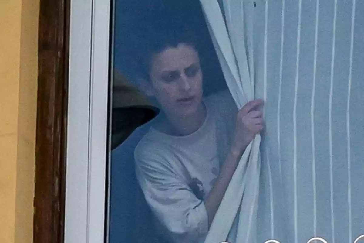 Persona asomándose por una ventana mientras sostiene una cortina blanca con rayas verticales Persona asomándose por una ventana mientras sostiene una cortina blanca con rayas verticales
