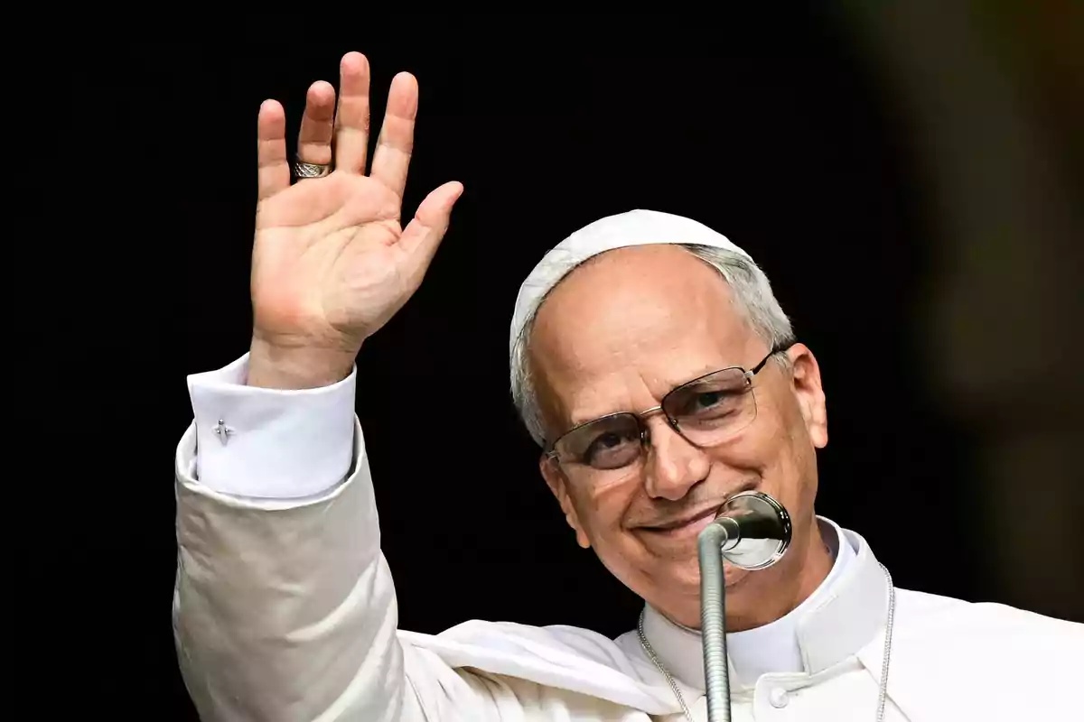 Hombre con vestimenta religiosa blanca y solideo saludando y sonriendo frente a un micrófono