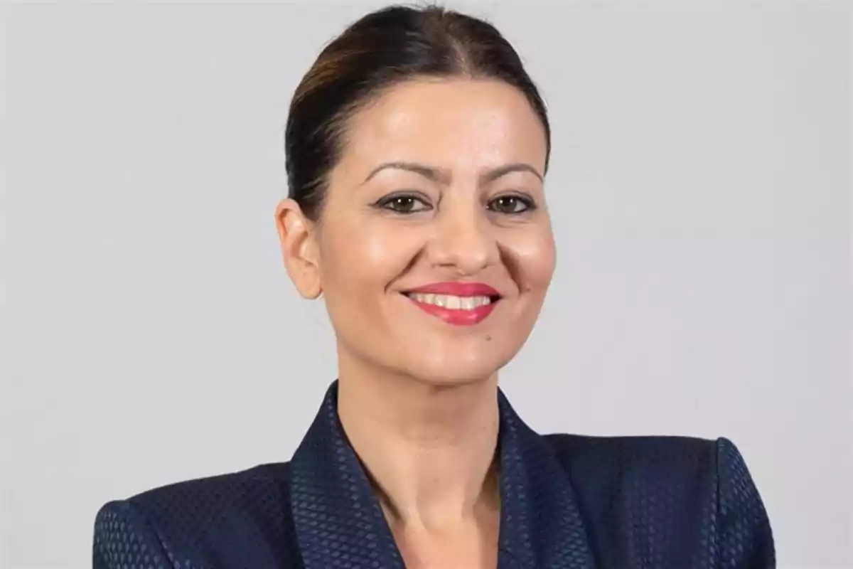 Mujer de cabello oscuro recogido con saco azul y sonrisa frente a fondo gris Mujer de cabello oscuro recogido con saco azul y sonrisa frente a fondo gris