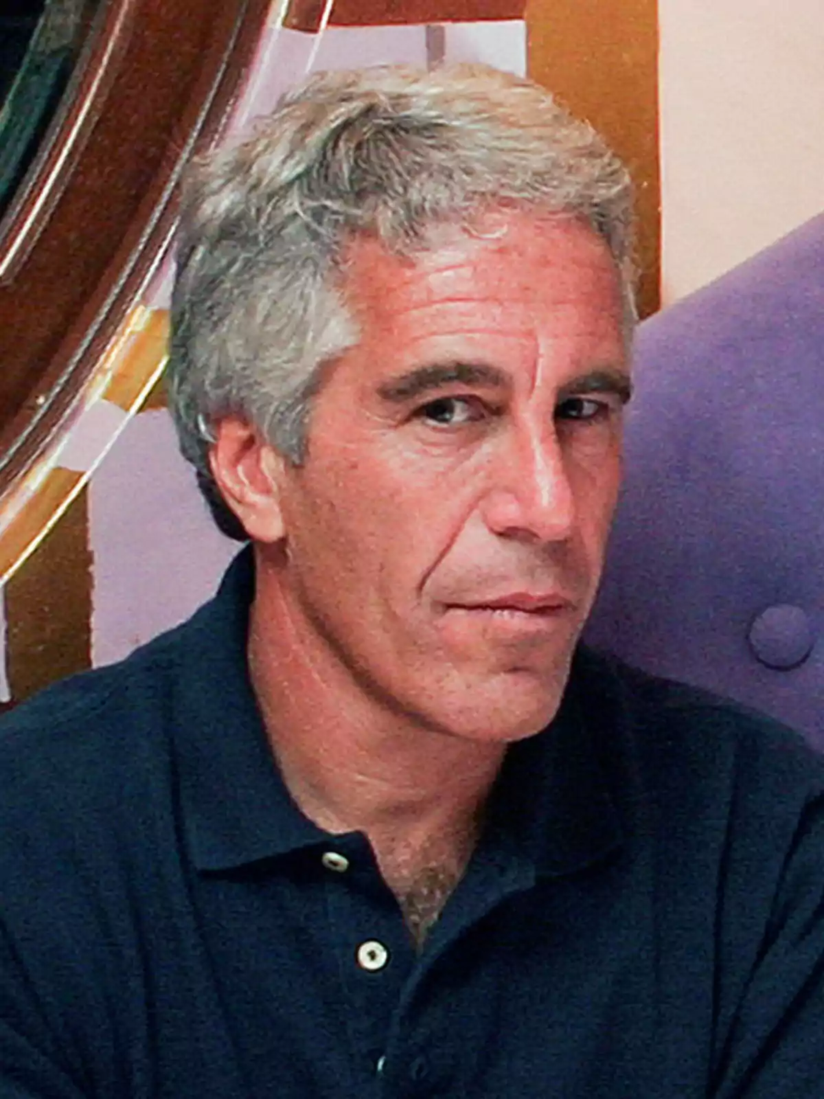 Hombre de cabello canoso y camisa polo oscura sentado frente a una pared colorida Hombre de cabello canoso y camisa polo oscura sentado frente a una pared colorida