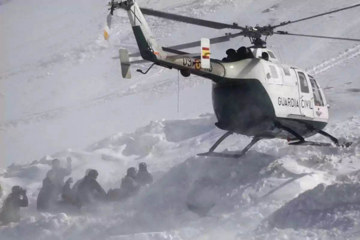 Helicóptero de la Guardia Civil aterrizando en una zona nevada mientras varias personas se protegen del viento y la nieve en el suelo Helicóptero de la Guardia Civil aterrizando en una zona nevada mientras varias personas se protegen del viento y la nieve en el suelo