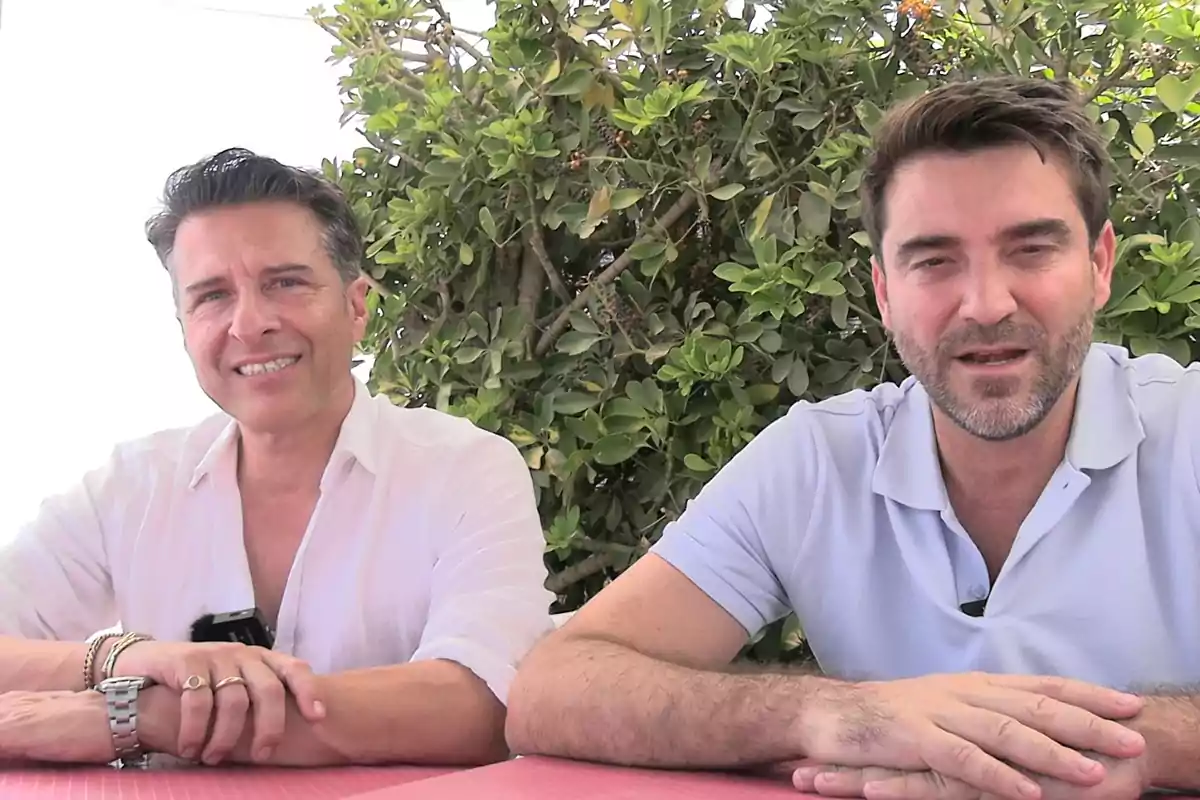 Dos hombres sentados en una mesa al aire libre con vegetación de fondo ambos sonríen y llevan camisas claras Dos hombres sentados en una mesa al aire libre con vegetación de fondo ambos sonríen y llevan camisas claras