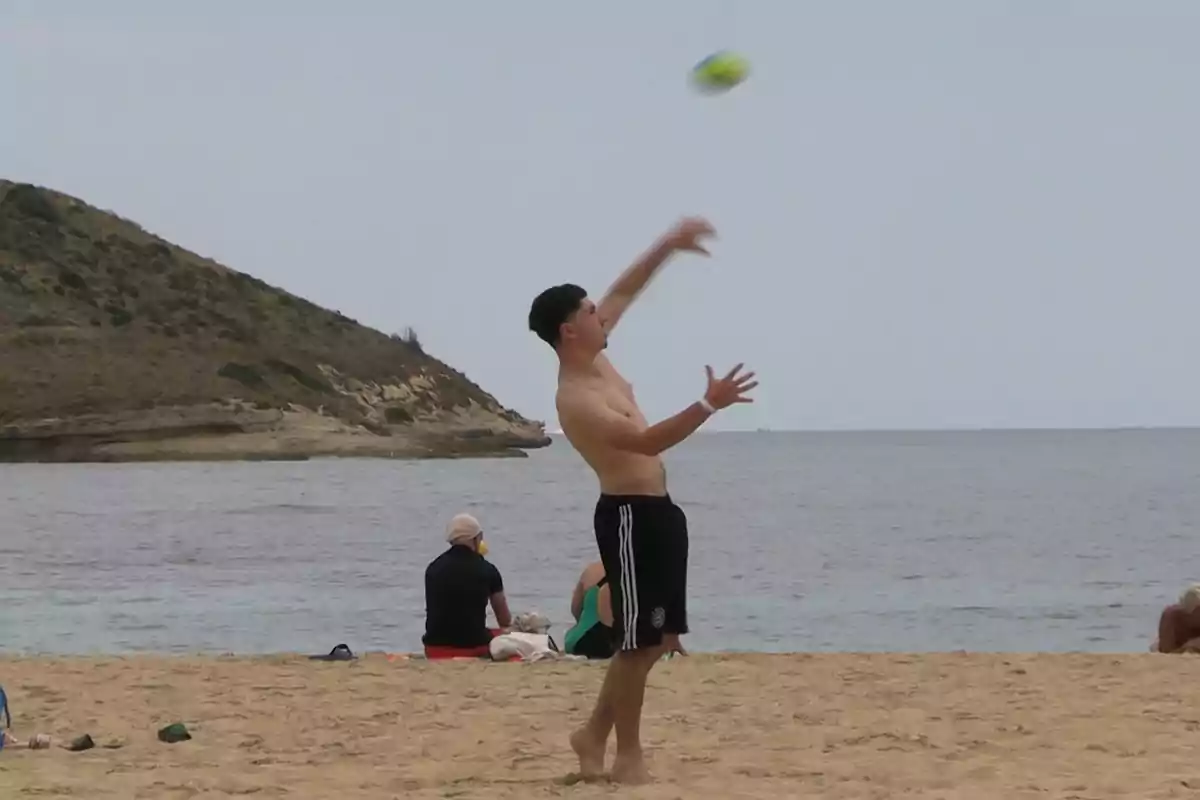 Joven lanzando una pelota en la playa con el mar y una colina al fondo mientras otras personas descansan en la arena Joven lanzando una pelota en la playa con el mar y una colina al fondo mientras otras personas descansan en la arena