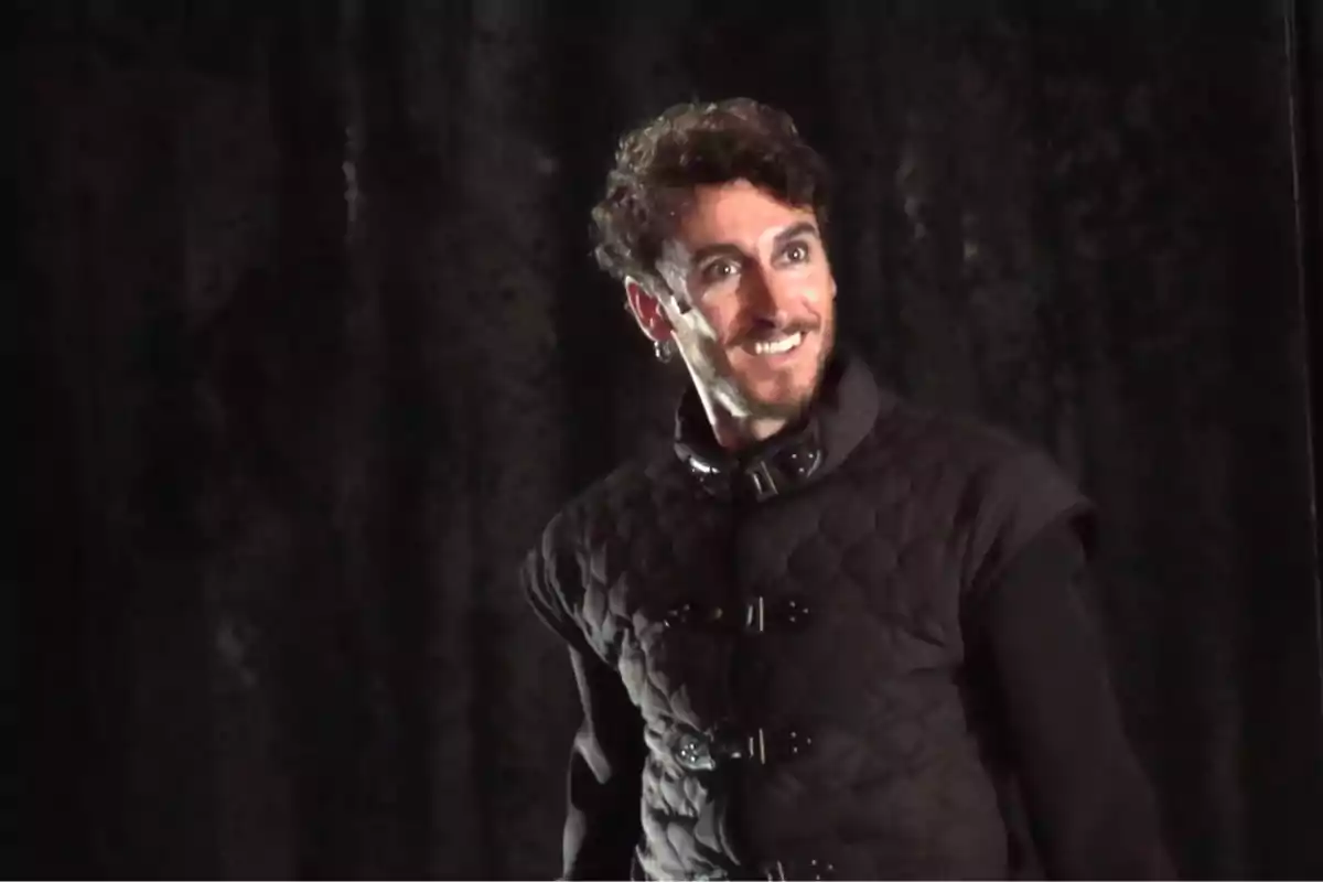 Hombre con chaleco oscuro y expresión sonriente en un escenario con fondo negro Hombre con chaleco oscuro y expresión sonriente en un escenario con fondo negro