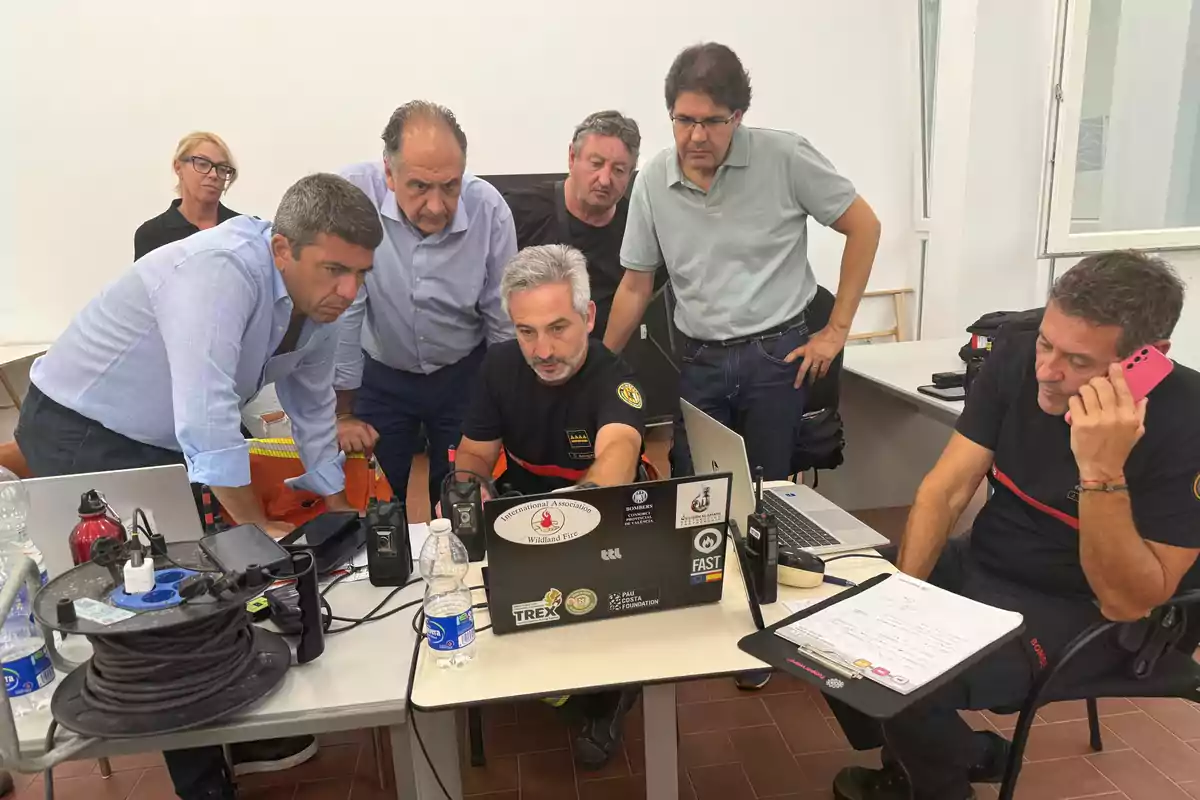 Un grupo de personas observa atentamente la pantalla de una computadora portátil en una sala de reuniones mientras un hombre habla por teléfono y hay varios equipos electrónicos y documentos sobre la mesa. Un grupo de personas observa atentamente la pantalla de una computadora portátil en una sala de reuniones mientras un hombre habla por teléfono y hay varios equipos electrónicos y documentos sobre la mesa.