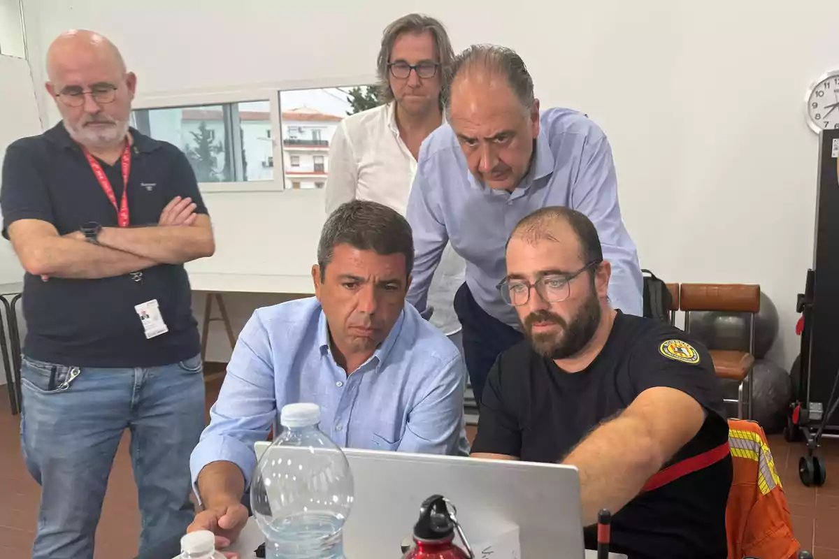 Cinco hombres observan atentamente la pantalla de una computadora portátil en una sala iluminada, mostrando concentración y trabajo en equipo.