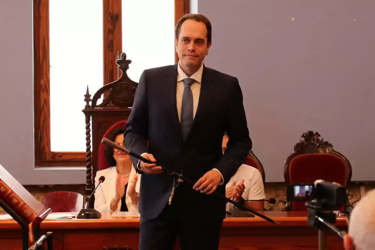 Hombre con traje oscuro y corbata azul de pie en una sala de reuniones sosteniendo un bastón mientras varias personas sentadas aplauden al fondo Hombre con traje oscuro y corbata azul de pie en una sala de reuniones sosteniendo un bastón mientras varias personas sentadas aplauden al fondo
