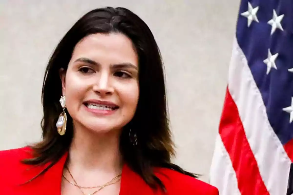 Mujer de cabello oscuro con saco rojo y aretes grandes de perla de pie junto a una bandera de Estados Unidos Mujer de cabello oscuro con saco rojo y aretes grandes de perla de pie junto a una bandera de Estados Unidos