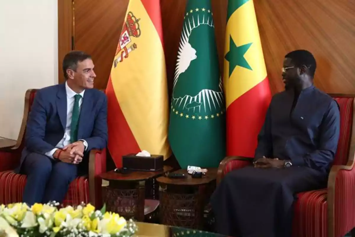 Dos hombres sentados en sillones conversan cordialmente con las banderas de España, la Unión Africana y Senegal detrás de ellos Dos hombres sentados en sillones conversan cordialmente con las banderas de España, la Unión Africana y Senegal detrás de ellos